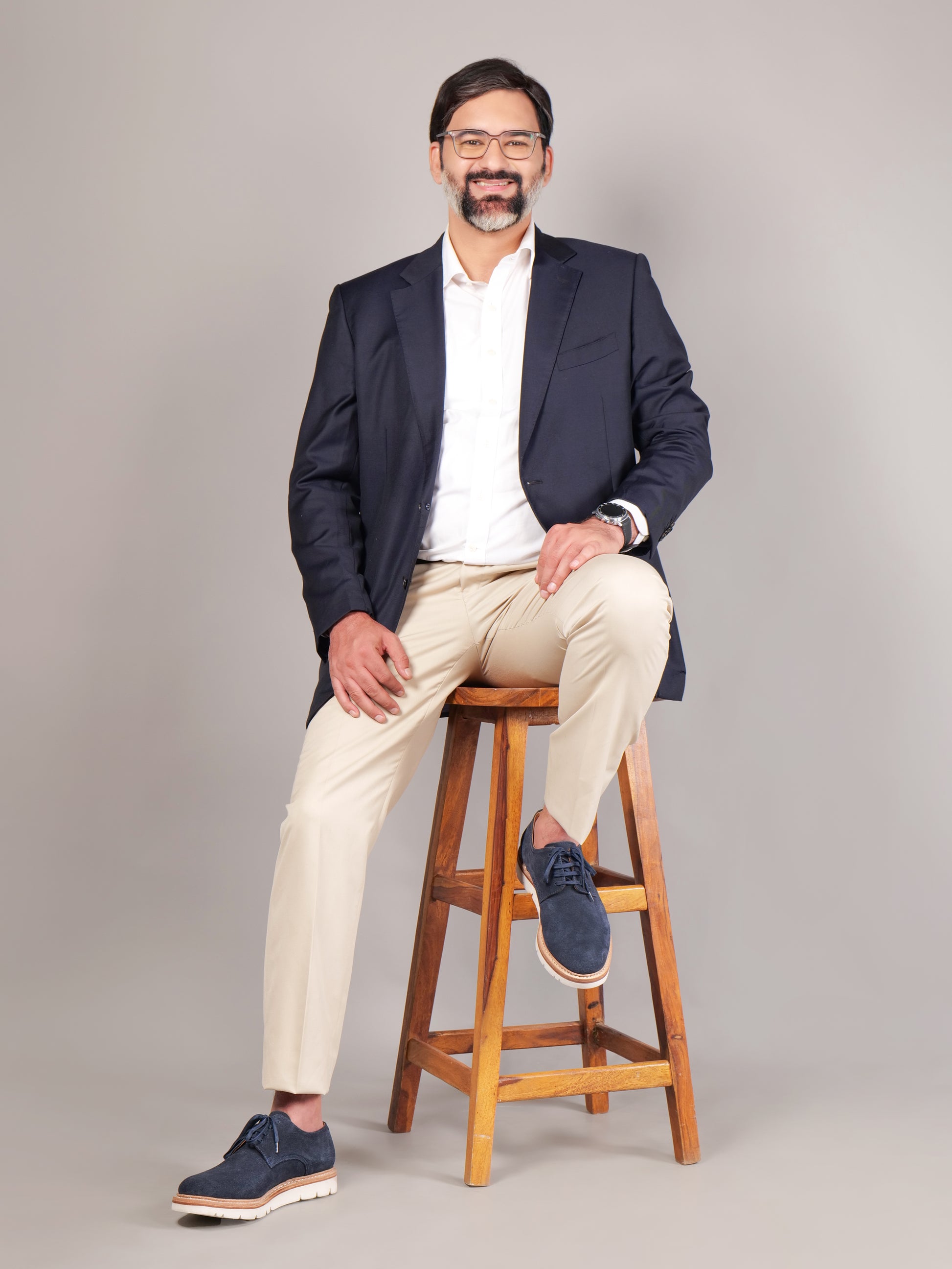 Male model wearing navy blue Blucher Suede shoes while seated on a stool against a beige background